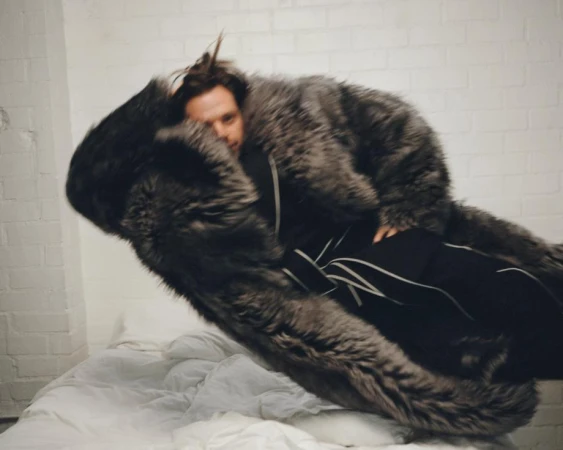 Person with dark hair in a gray fur coat sitting on a bed in front of a white brick wall, posing indoors with a neutral expression.