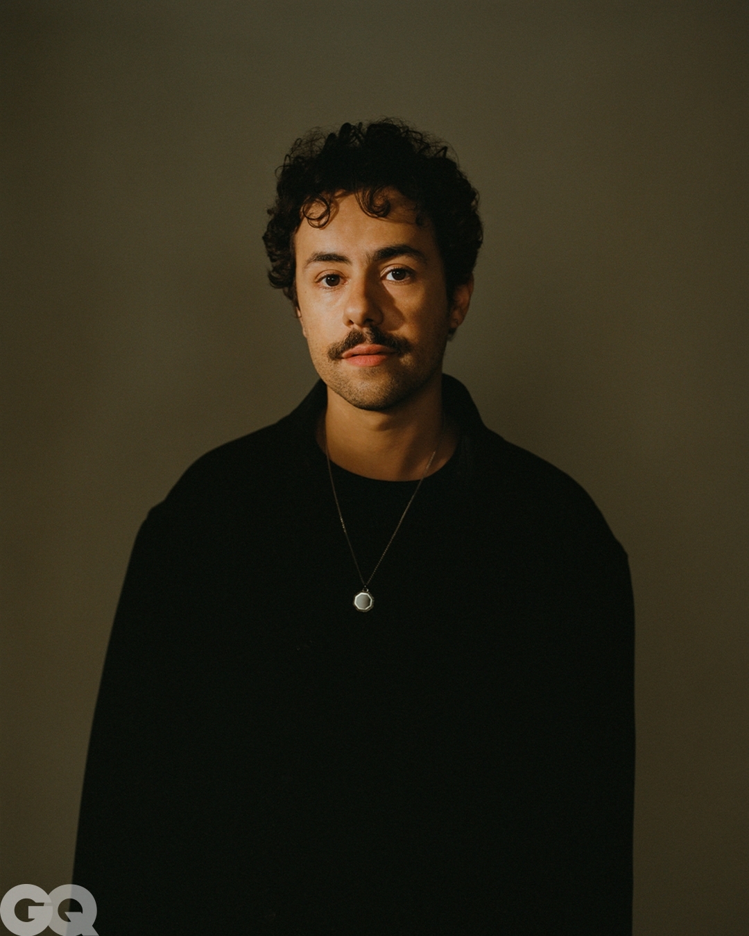 Portrait of a young adult male with dark curly hair and a mustache, wearing a black collared shirt and a necklace with a pendant.