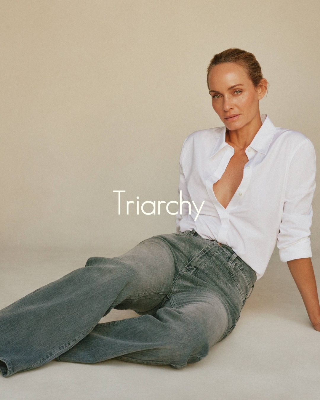 Adult woman in unbuttoned white shirt and grey jeans poses in a photo studio with tan backdrop, looking at the camera with a neutral expression.