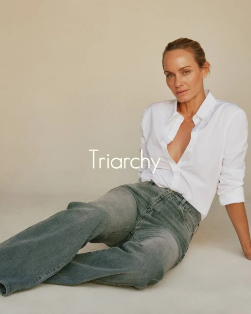 Adult woman in unbuttoned white shirt and grey jeans poses in a photo studio with tan backdrop, looking at the camera with a neutral expression.