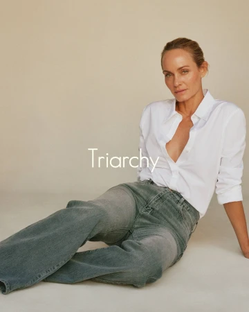 Adult woman in unbuttoned white shirt and grey jeans poses in a photo studio with tan backdrop, looking at the camera with a neutral expression.