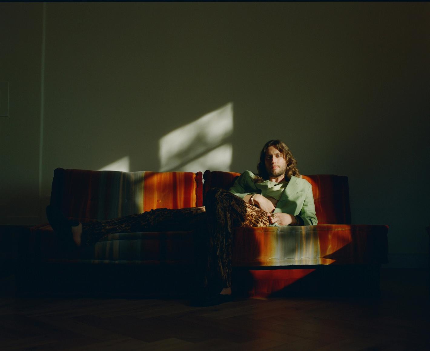 Man with long hair sits on a colorful sofa in a sunlit room, wearing a green jacket and patterned pants in a relaxed pose.