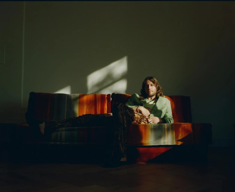 Man with long hair sits on a colorful sofa in a sunlit room, wearing a green jacket and patterned pants in a relaxed pose.