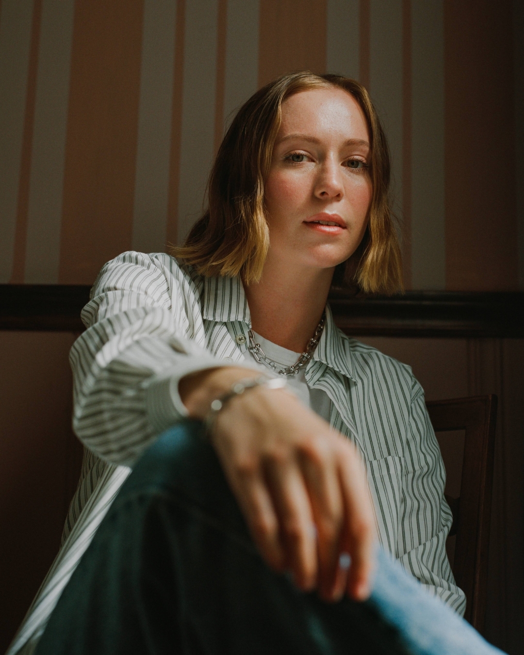 Portrait of a young woman with short blonde hair wearing a striped shirt and jeans, seated in an indoor setting with soft lighting.