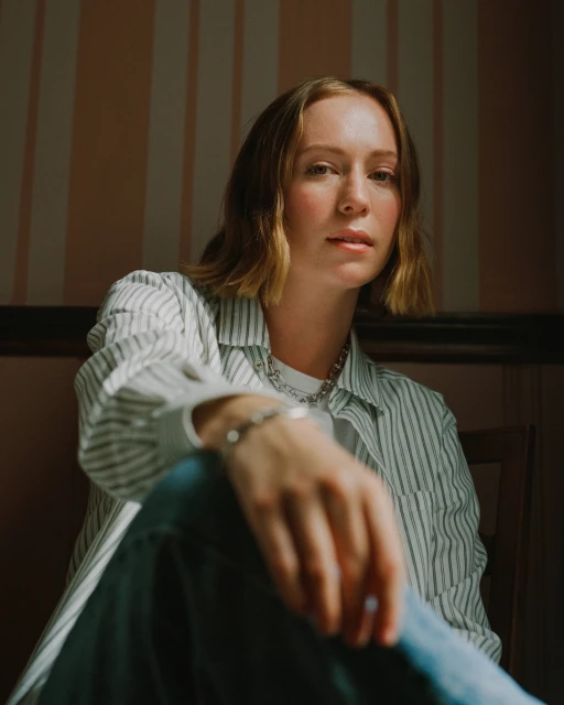 Portrait of a young woman with short blonde hair wearing a striped shirt and jeans, seated in an indoor setting with soft lighting.
