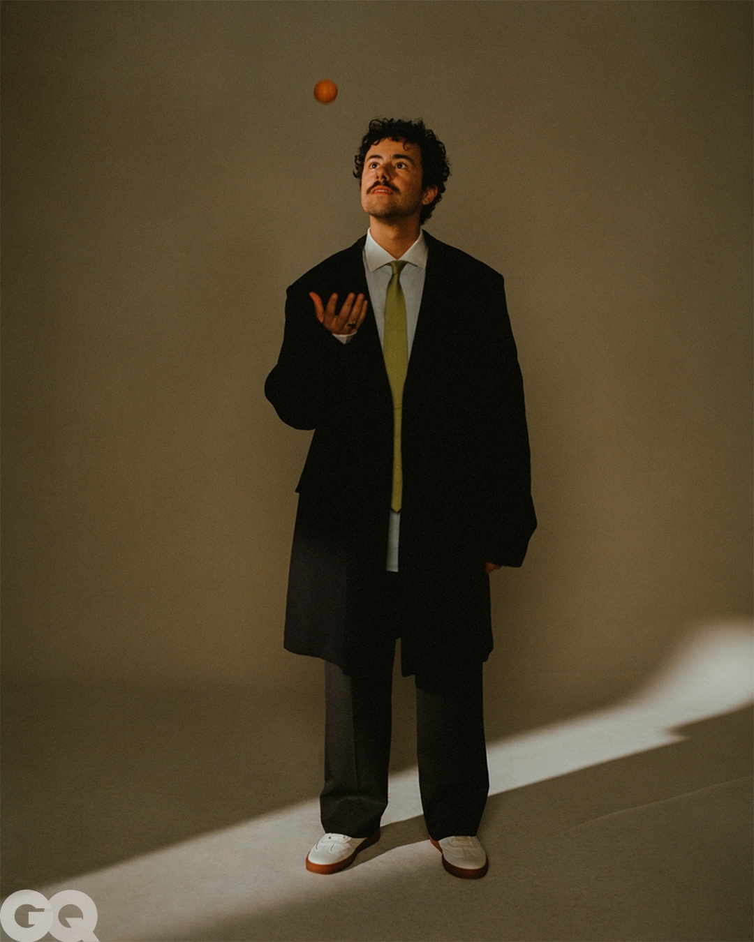 Adult male in black coat and pants stands in a neutral studio, juggling an orange in mid-air, photographed in a full shot.