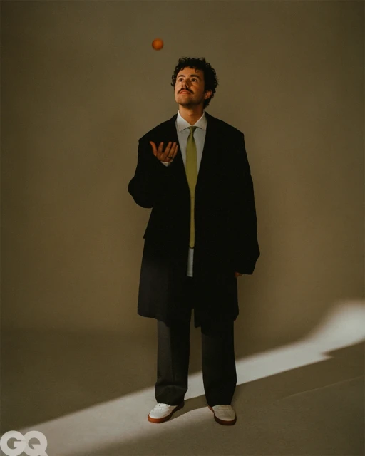 Adult male in black coat and pants stands in a neutral studio, juggling an orange in mid-air, photographed in a full shot.