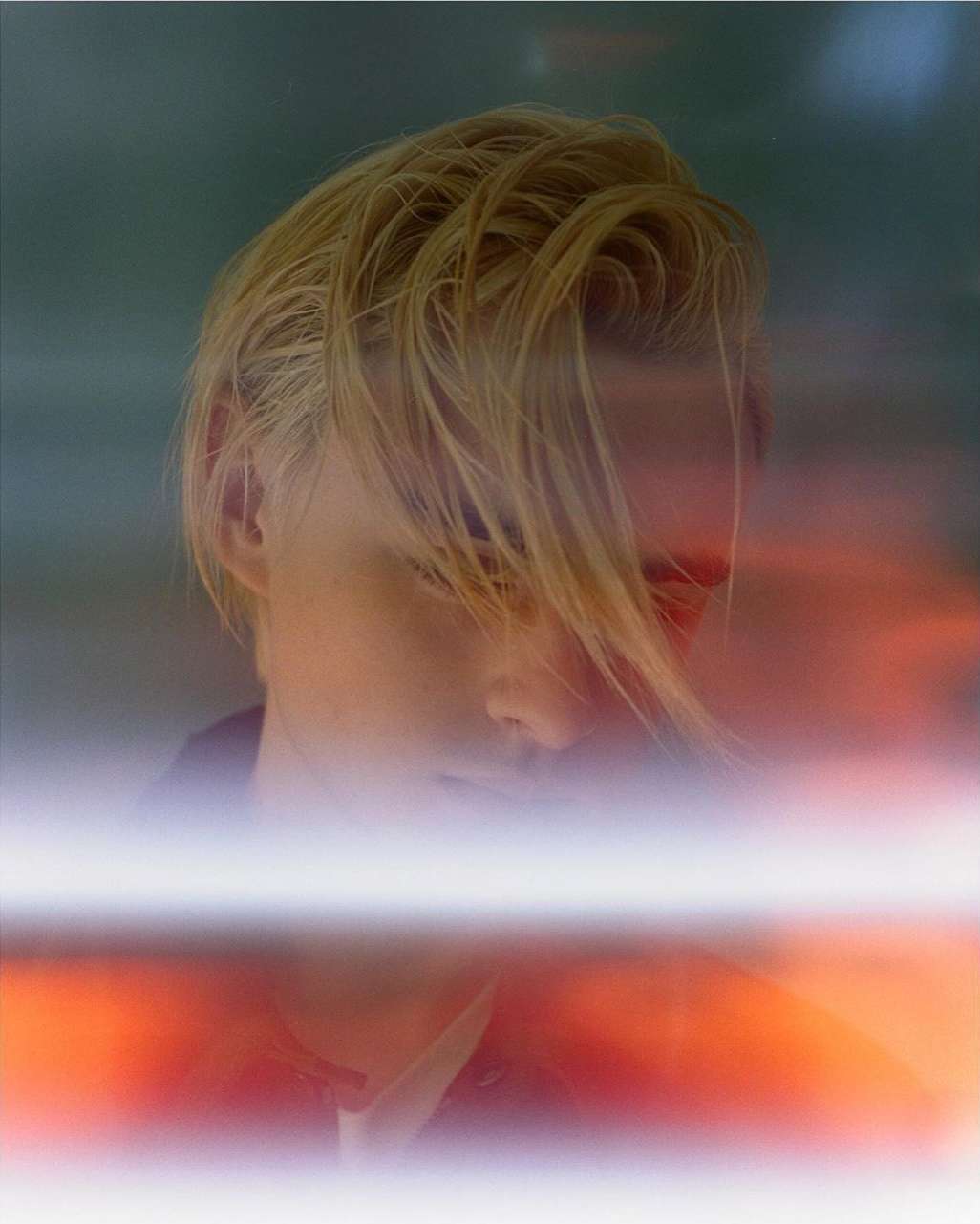 Portrait of a fair-skinned adult with blond hair, partially obscured by blurred orange and white light, set against a muted green background.