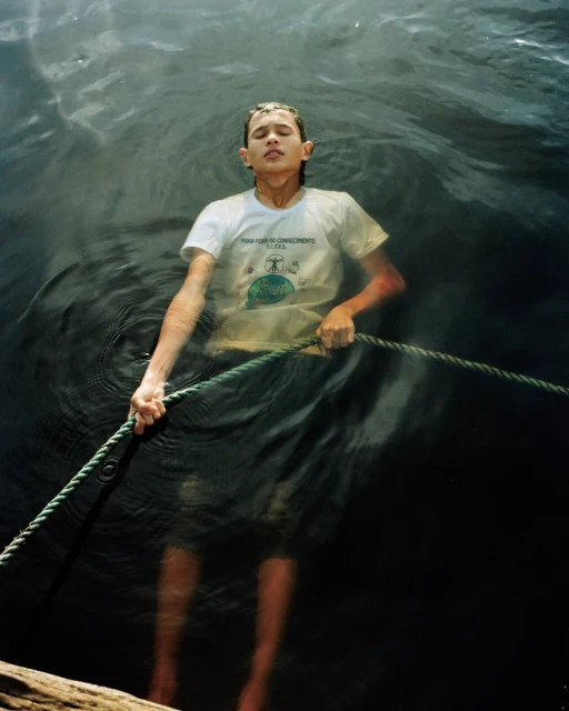 Teenager in water stands holding rope, wearing a white t-shirt with graphic, eyes closed, in an outdoor natural setting.