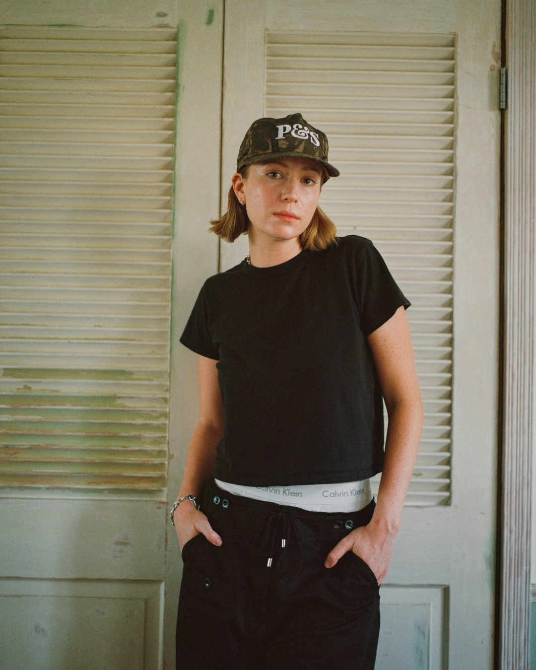 Adult woman in casual black top and bottoms, wearing a baseball cap, standing indoors against a white shuttered door.