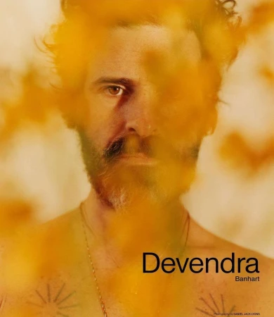 Portrait of Devendra Banhart with a beard and tattoos, partially obscured by out-of-focus yellow foliage in the foreground.