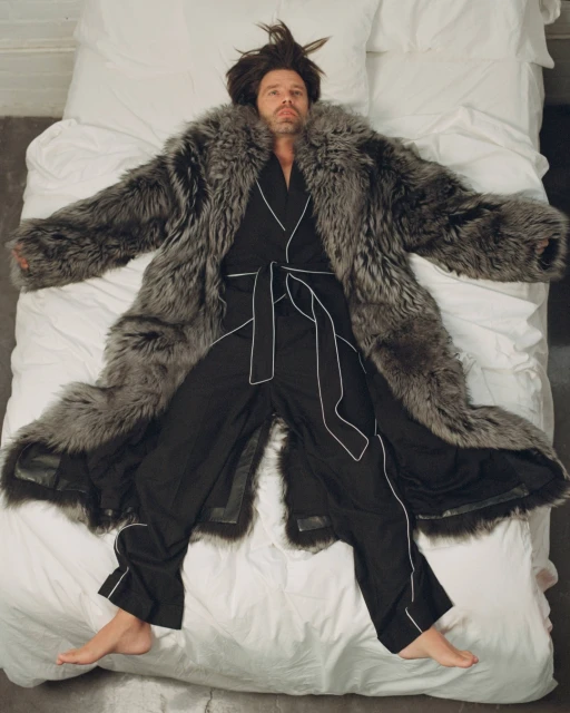 Adult male lies on a white bed wearing a fur coat and black pajamas with white piping, viewed from an overhead angle in an indoor setting.