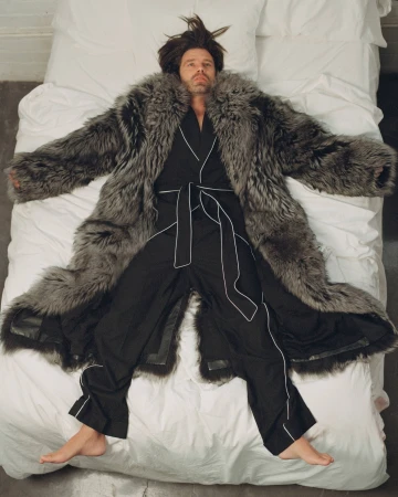 Adult male lies on a white bed wearing a fur coat and black pajamas with white piping, viewed from an overhead angle in an indoor setting.