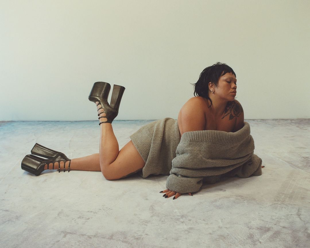 Woman lying on the floor wearing an off-the-shoulder sweater and platform heels, with a seamless white backdrop and minimalist aesthetic.