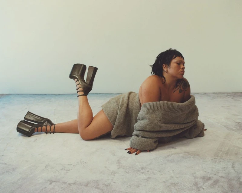 Woman lying on the floor wearing an off-the-shoulder sweater and platform heels, with a seamless white backdrop and minimalist aesthetic.