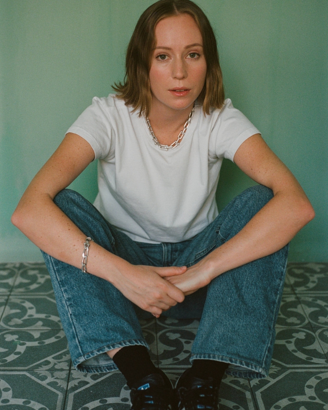 Woman in white t-shirt and blue jeans sitting on patterned tiled floor against green wall