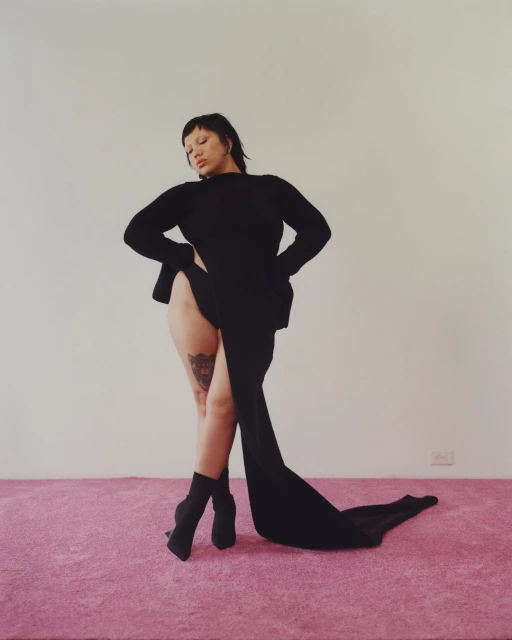 An adult female poses indoors wearing a black dress and boots, standing on a pink carpet against a light wall, in a medium shot.