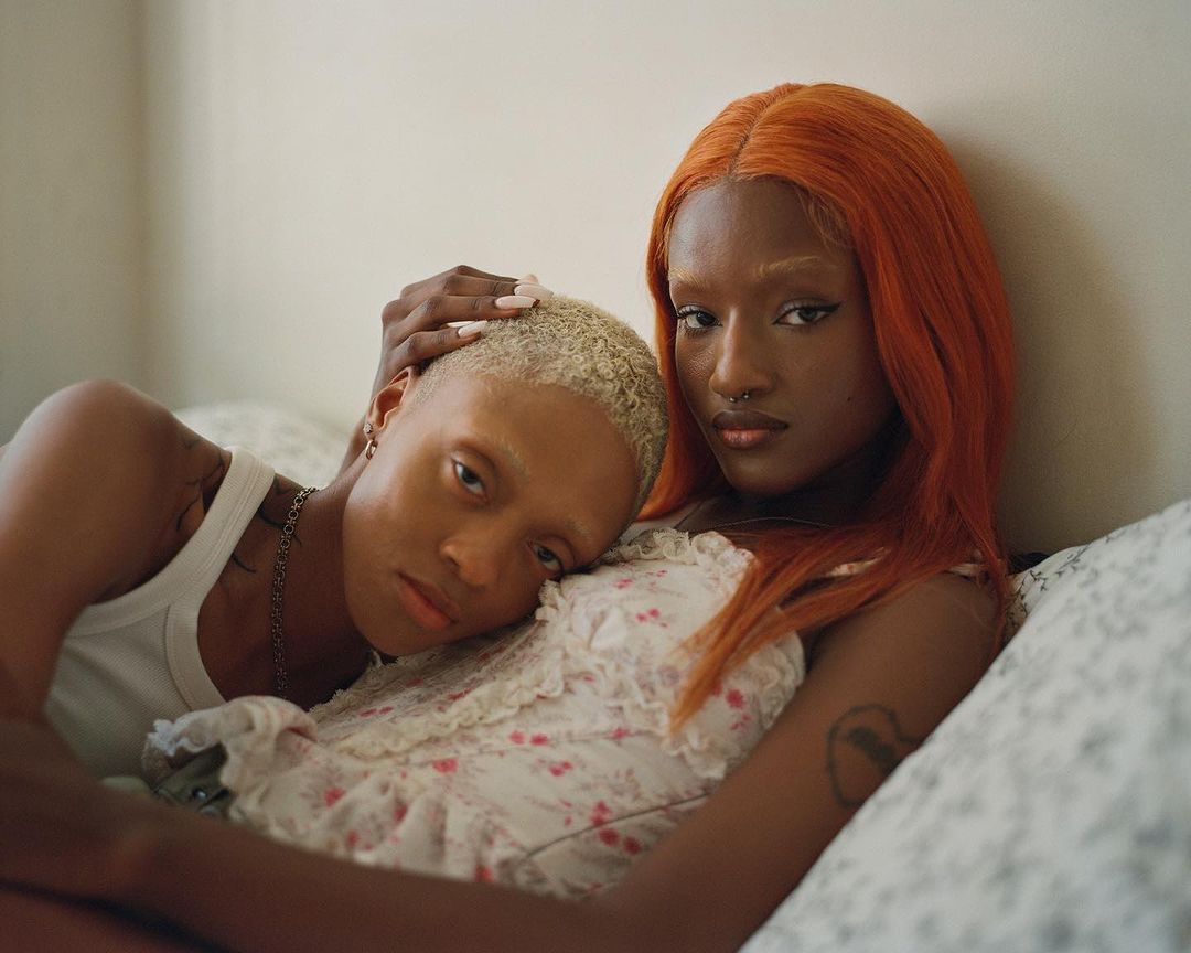 Two women resting indoors, one with bright orange hair and another with short blonde hair resting her head on her shoulder.