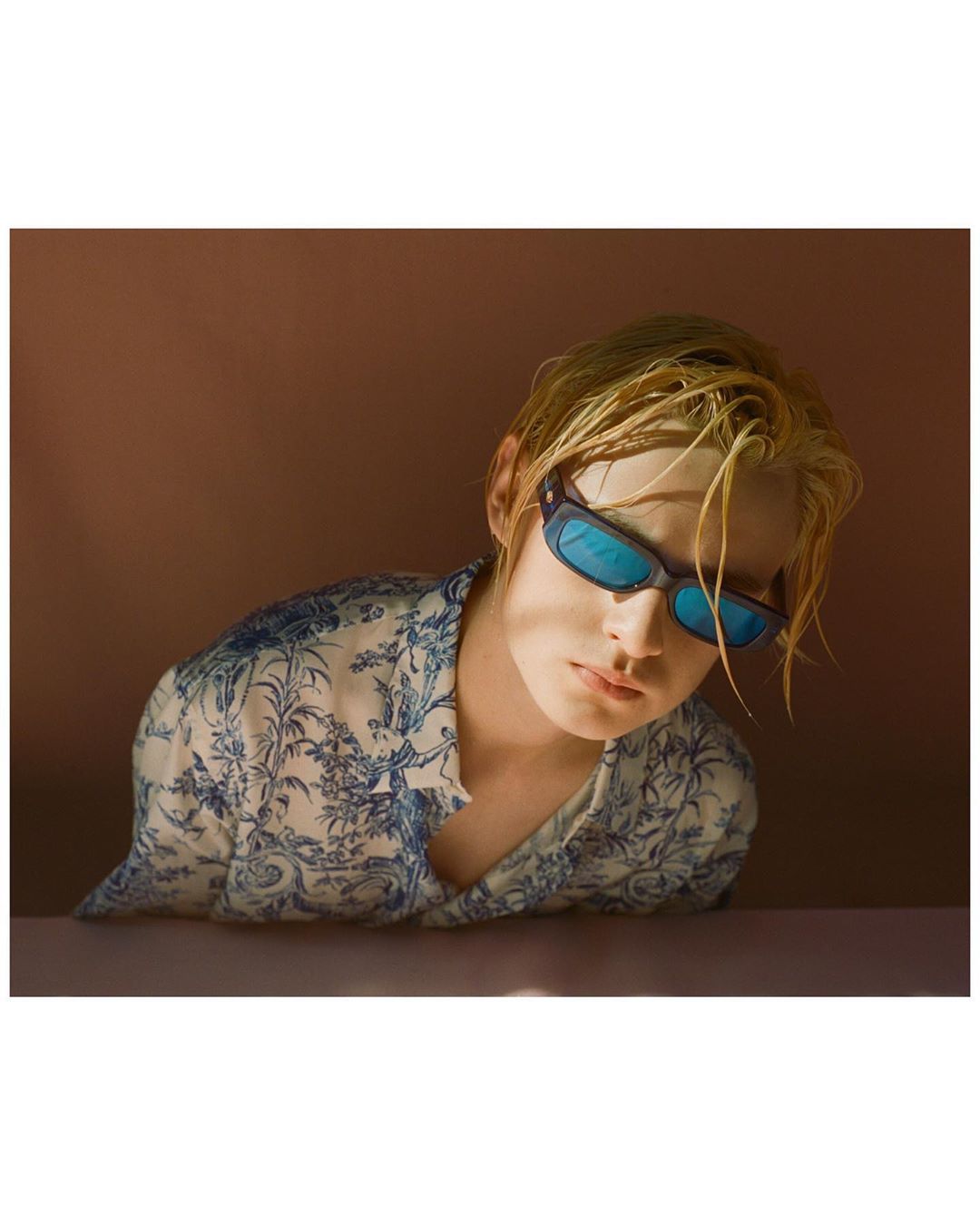 Portrait of a young adult with blonde hair and blue sunglasses wearing a floral shirt against a brown backdrop, eye-level shot.