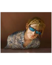 Young person with wet blonde hair wearing blue sunglasses and a blue-and-white patterned shirt against a warm brown background