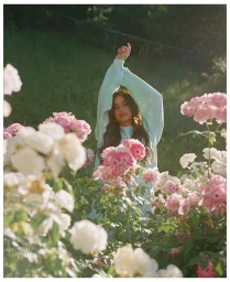 A young woman wearing a light blue sweatshirt poses amidst a lush garden of blooming pink and white roses.