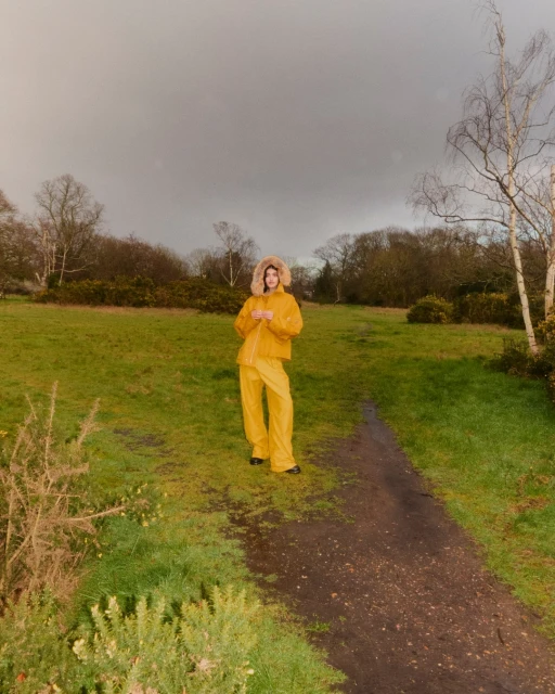 Woman in yellow hooded raincoat and pants stands on a grassy path in a park setting on a cloudy day, looking at the camera.