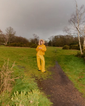 Woman in yellow hooded raincoat and pants stands on a grassy path in a park setting on a cloudy day, looking at the camera.