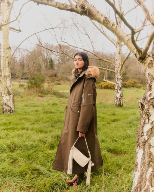 A woman in a fur-trimmed parka poses with a purse in a grassy field with bare trees. Autumn colors, overcast day.