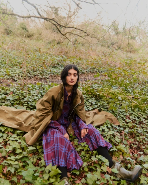 Adult woman in green coat and plaid dress sits outdoors on a bed of ivy, with trees and light sky visible in the background.