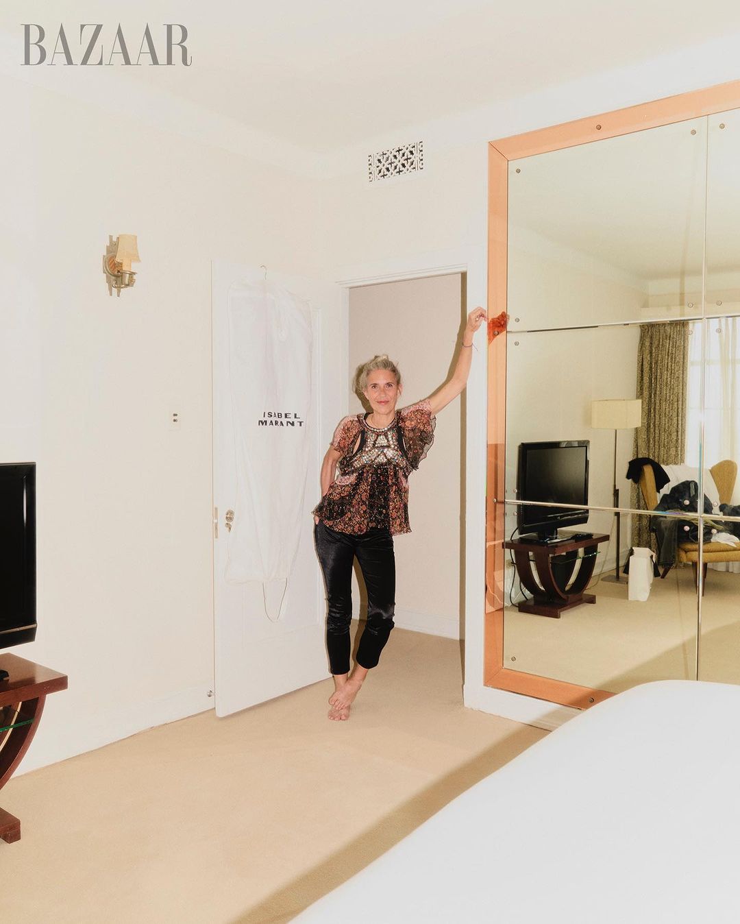 An adult woman in a patterned blouse and black pants poses inside a hotel room with a large mirror. Interior fashion shot.