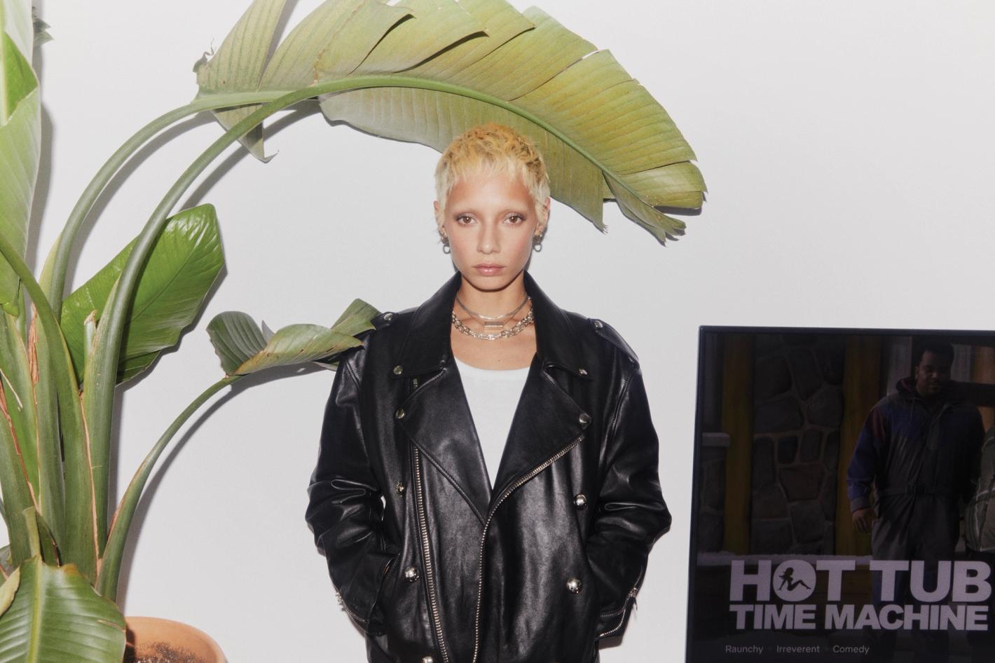 Woman with short blonde hair in a black leather jacket posing indoors with a large green plant and Hot Tub Time Machine poster.