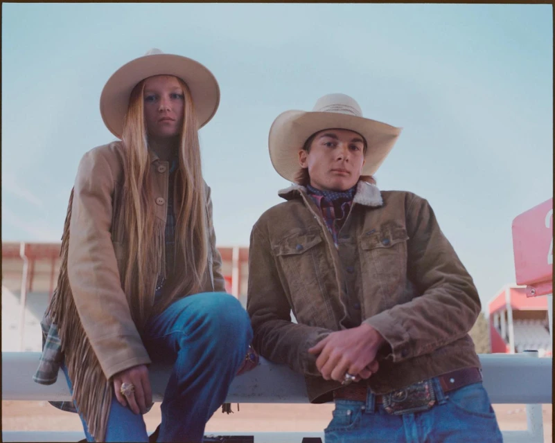 Two people in cowboy hats and jackets pose outdoors. The woman wears a fringed jacket, and the man has a denim jacket with a large belt buckle.