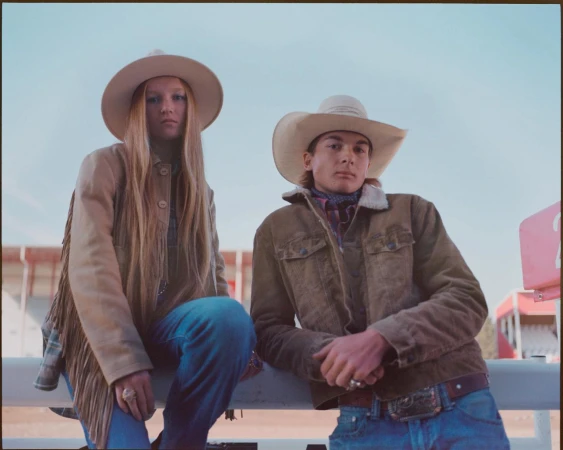 Two people in cowboy hats and jackets pose outdoors. The woman wears a fringed jacket, and the man has a denim jacket with a large belt buckle.