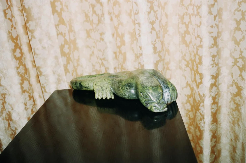 Green serpentine alligator sculpture on a dark wood table with patterned cream curtains behind it. Interior setting with neutral tones.