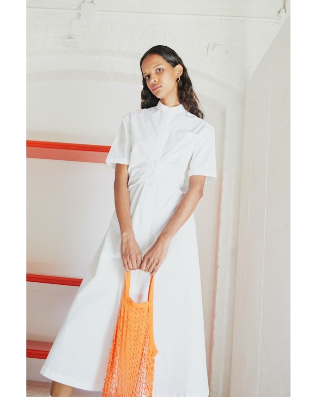 A woman in a white dress stands indoors holding an orange net bag, near an orange shelf and a white wall with an arched ceiling.
