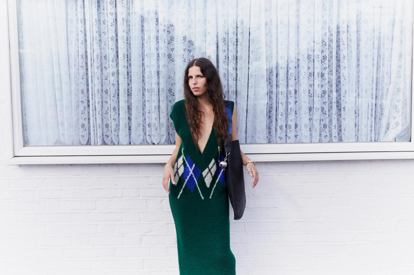 Adult female in a deep green knitted dress with a blue and white argyle pattern, posing outdoors against a white brick wall.
