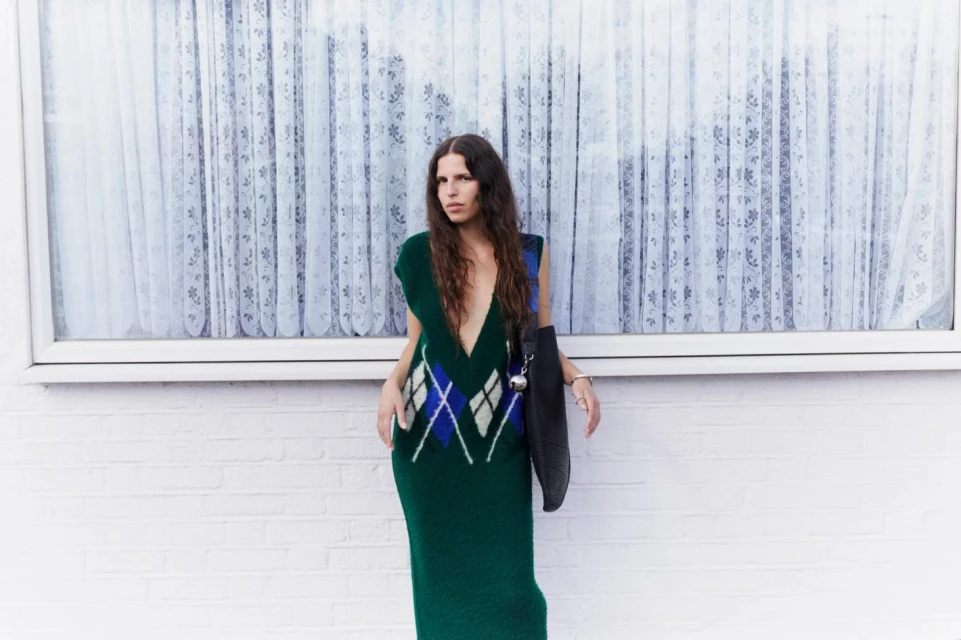 Adult female in a deep green knitted dress with a blue and white argyle pattern, posing outdoors against a white brick wall.
