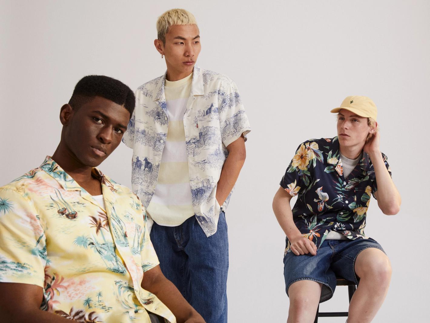 Three young adult men pose in a light studio setting. One man sits on a stool while the others stand behind him, wearing tropical patterned shirts.