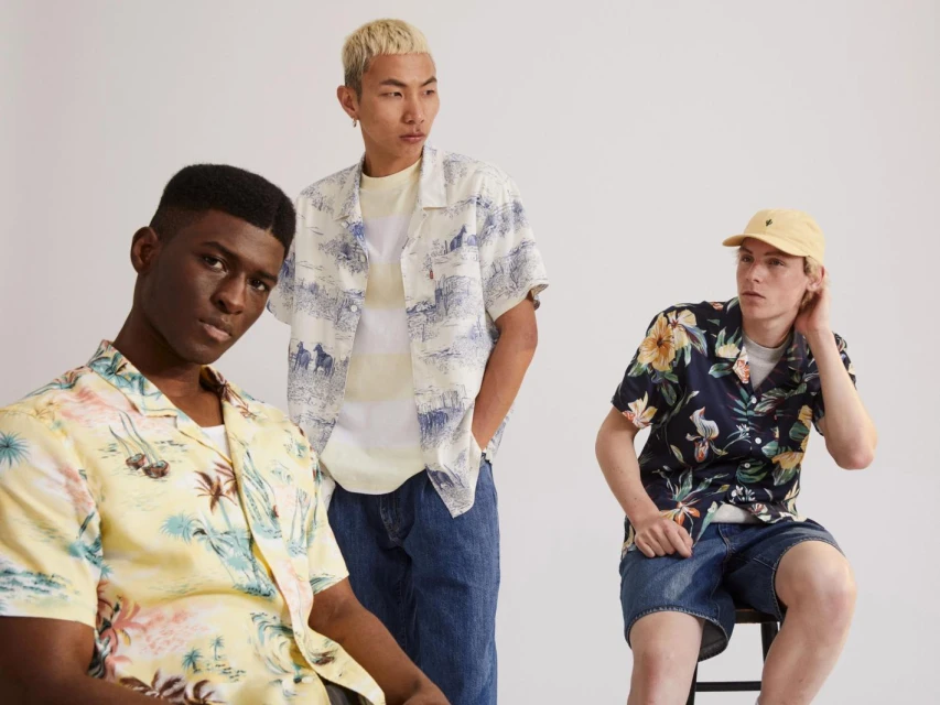 Three young adult men pose in a light studio setting. One man sits on a stool while the others stand behind him, wearing tropical patterned shirts.