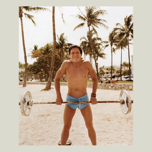 Shirtless mature man lifting barbell on beach surrounded by palm trees, bright daylight, fitness concept, weight training and outdoors.