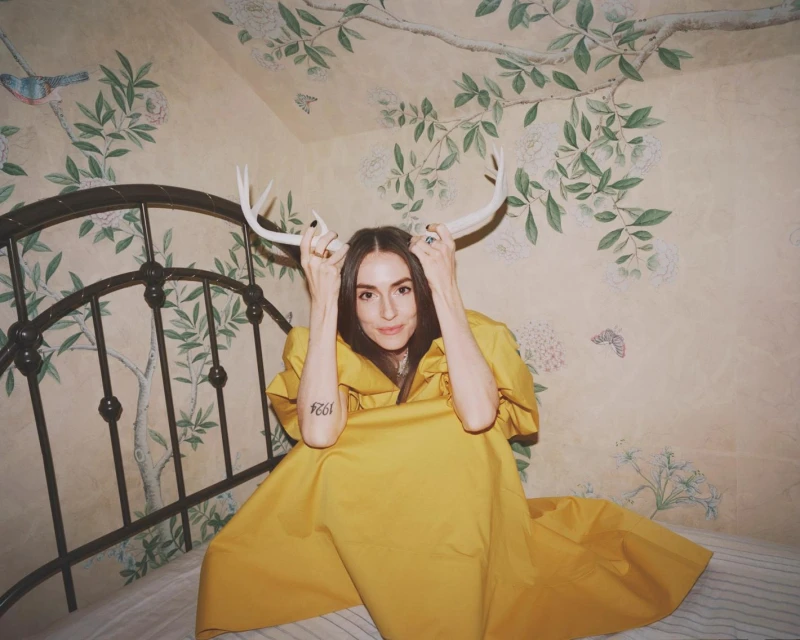 Woman in yellow dress sits on bed holding antlers in floral wallpaper room; indoor portrait with soft light and eye-level angle.