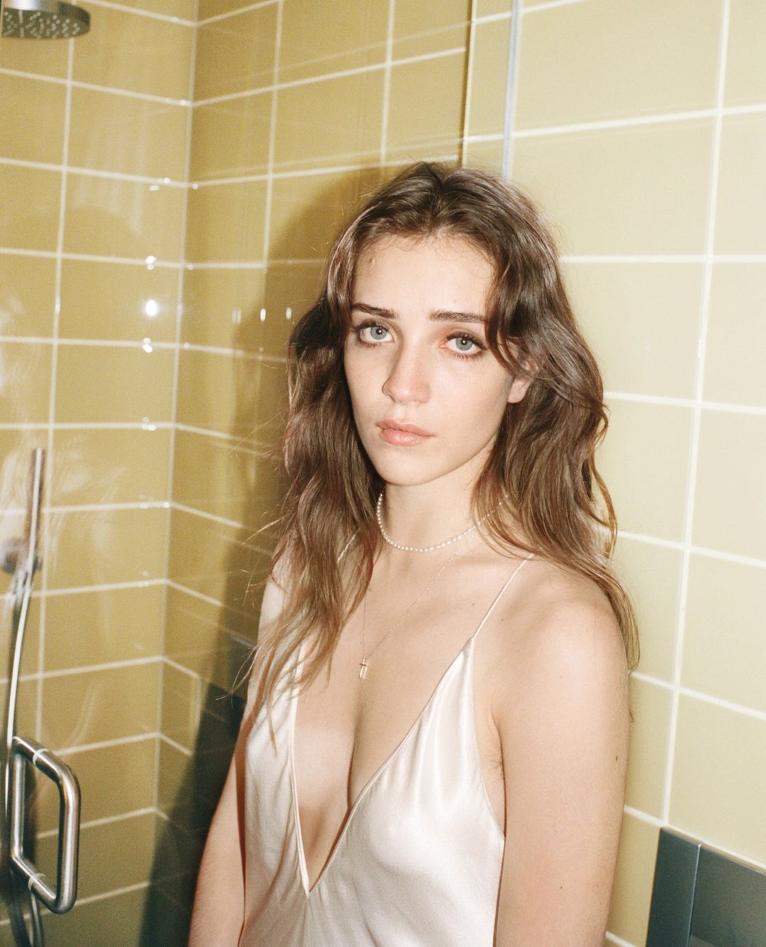 Close-up portrait of a young woman with long brown hair in a slip dress, posing indoors against a tiled wall with a showerhead.