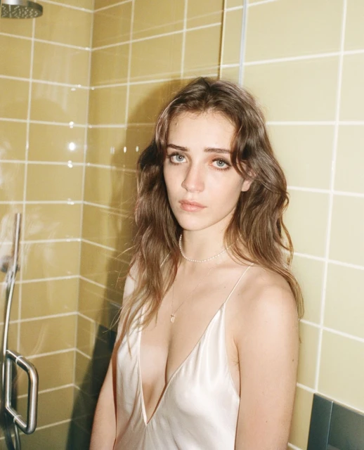 Close-up portrait of a young woman with long brown hair in a slip dress, posing indoors against a tiled wall with a showerhead.