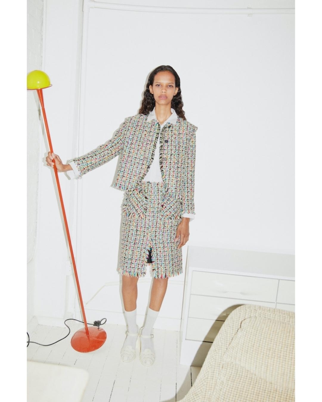 Fashion editorial shot of an adult female model in a tweed skirt and jacket posing in a studio with a floor lamp, white walls, and white furniture.