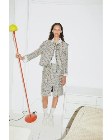 Fashion editorial shot of an adult female model in a tweed skirt and jacket posing in a studio with a floor lamp, white walls, and white furniture.