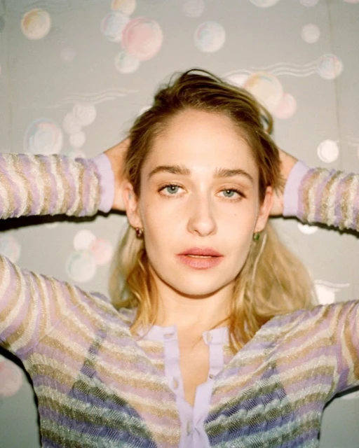 Blonde woman with hands behind her head poses in a pastel-striped shirt with a colorful bubble pattern in the background.