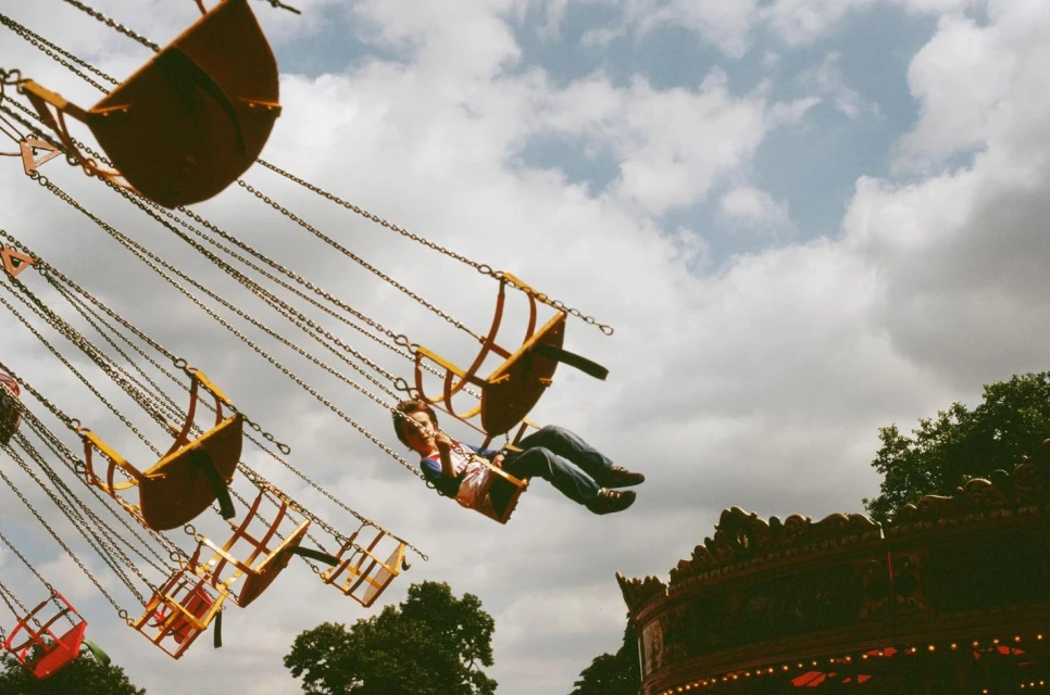 A person enjoys a fairground ride with yellow seats suspended by chains against a partly cloudy sky, seen from a low angle.