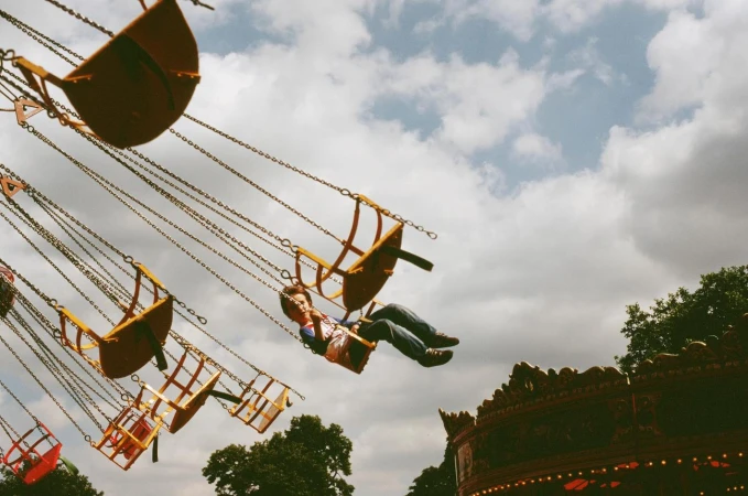 A person enjoys a fairground ride with yellow seats suspended by chains against a partly cloudy sky, seen from a low angle.