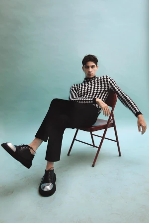 Man seated on a red folding chair wearing a black and white checkerboard sweater and silver-capped black shoes against a light blue backdrop.