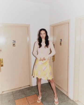 Model wearing a patterned yellow skirt and long-sleeve top standing in a hallway between doors labeled 2A and 2B.